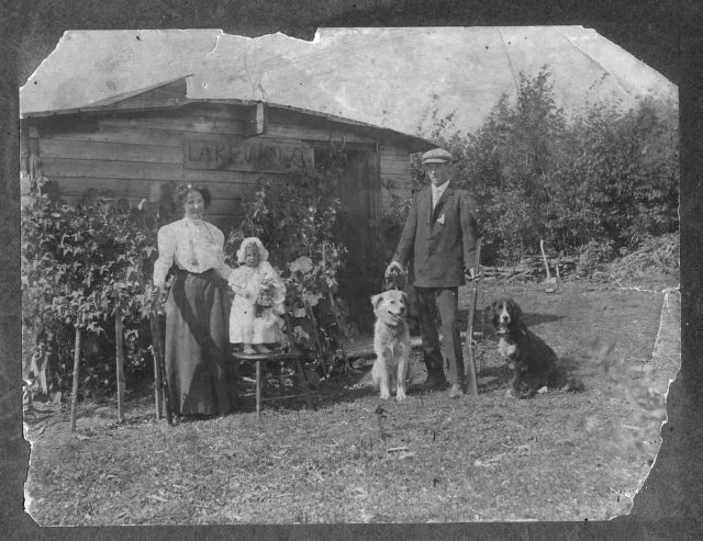 A family with a young child and two dogs standing outside their homestead