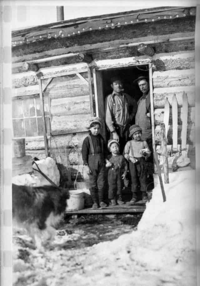 Two men and three children standing in the doorway of a log cabin home