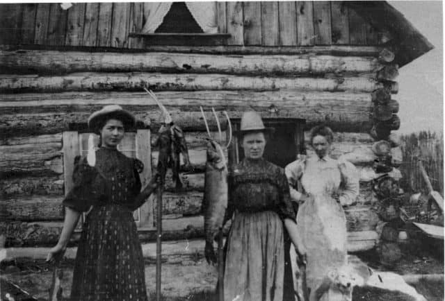Three woman holding fish on pitchforks standing in front of a log cabin home