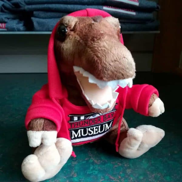 Dinosaur Plushie wearing a red sweater with the Thunder Bay Museum logo on it