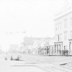 Old faded photograph of buildings along a street. There's some people standing around in small groups.