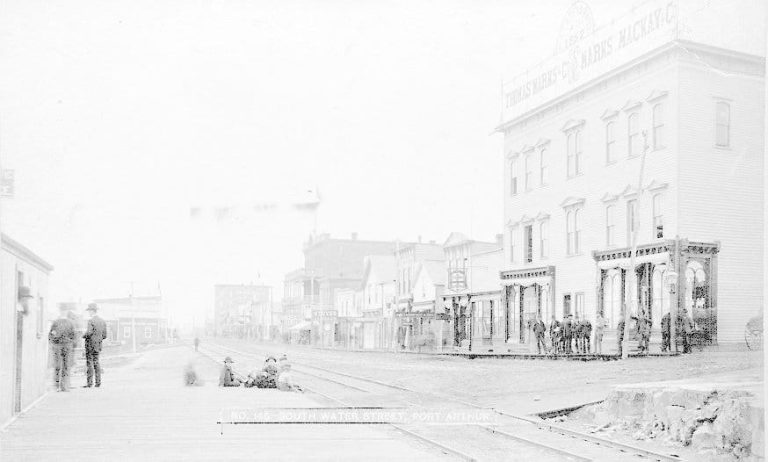 Old faded photograph of buildings along a street. There's some people standing around in small groups.