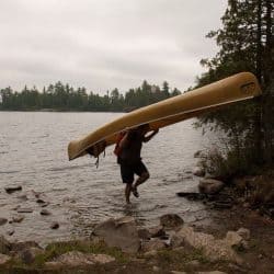 Michael deJong carrying a canoe over his head at the edge of a lake.