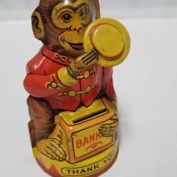 Tin monkey holding a yellow hat one hand with a red vest on. The monkey is sitting on a box that says Bank and there's a slot to put coins in.