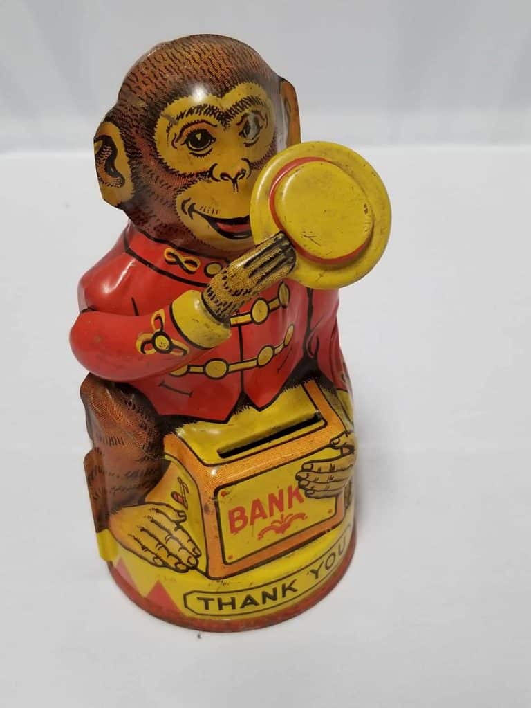 Tin monkey holding a yellow hat one hand with a red vest on. The monkey is sitting on a box that says Bank and there's a slot to put coins in.