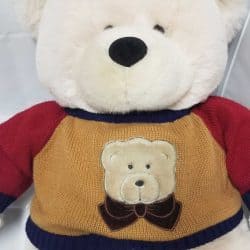 Closeup of a white teddy bear and its gold, red, and blue knit sweater featuring a bear head patch on the front of the sweater.